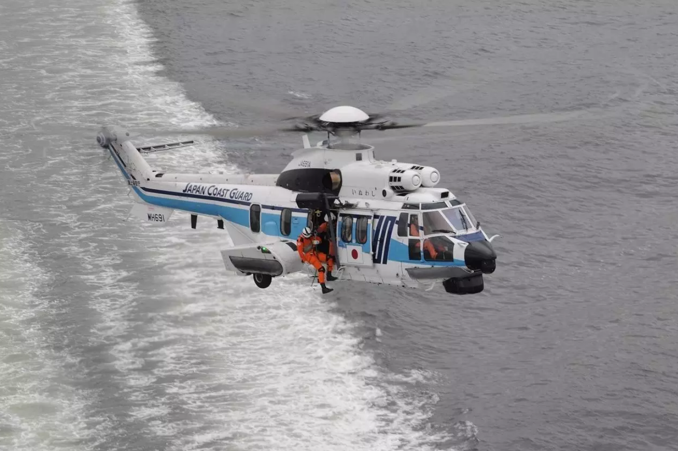 A Japan Coast Guard H225 helicopter is seen flying above gray waters. The white aircraft has a thin navy blue and thick light blue stripe painted on its side, as well as 