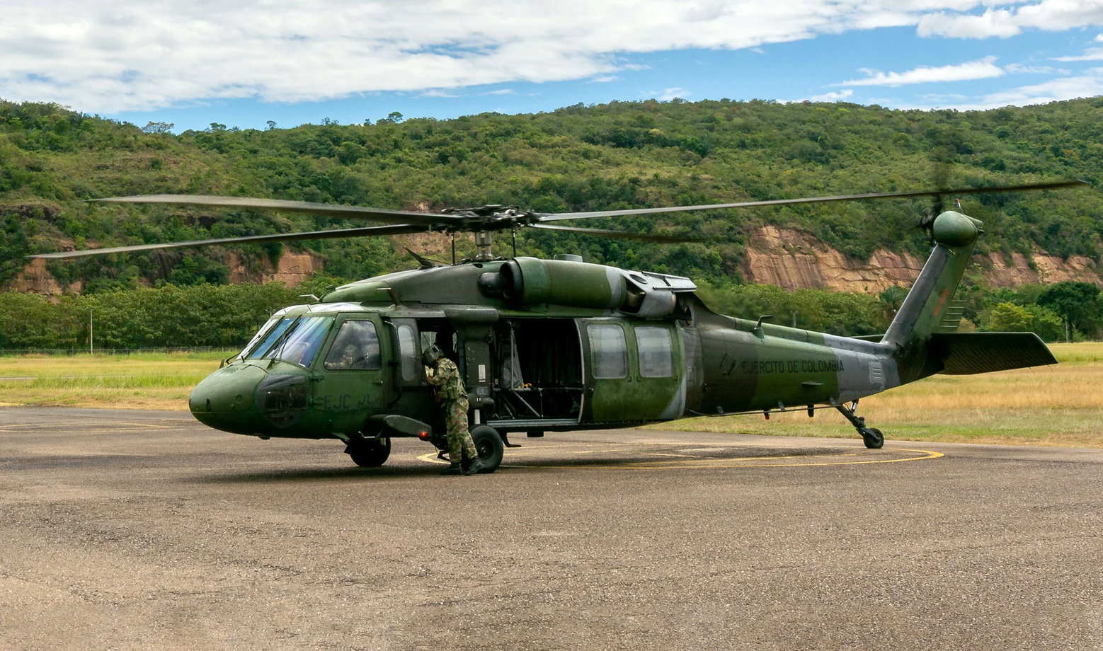 A Colombian Army Black Hawk helicopter is seen grounded in a large open cemented field. The helicopter is painted various hues of dark green as camouflage. A soldier in camouflage is seen standing by the cockpit. 