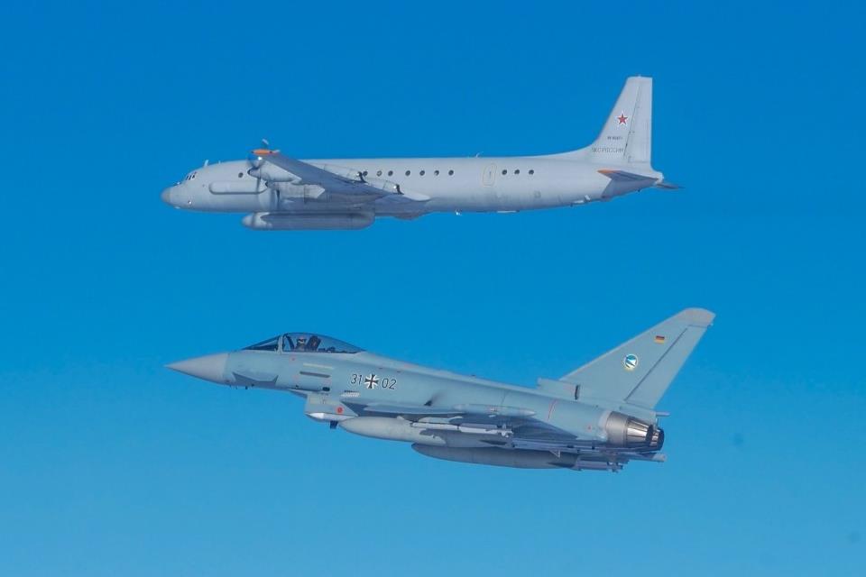 A German Eurofighter flies below another aircraft. Both planes are painted gray, with the Eurofighter having a darker tone. The background is a clear blue sky.