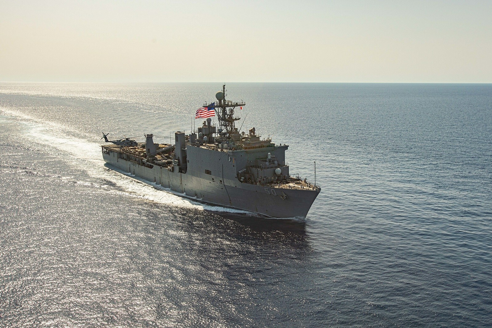 The dock landing ship USS Carter Hall (LSD-50) is seen sailing in calm waters. The sun, out of frame, shines a birght yellow-orange hue on the waters and the ship. The ship is painted gray, and has the US flag flying at full mast.