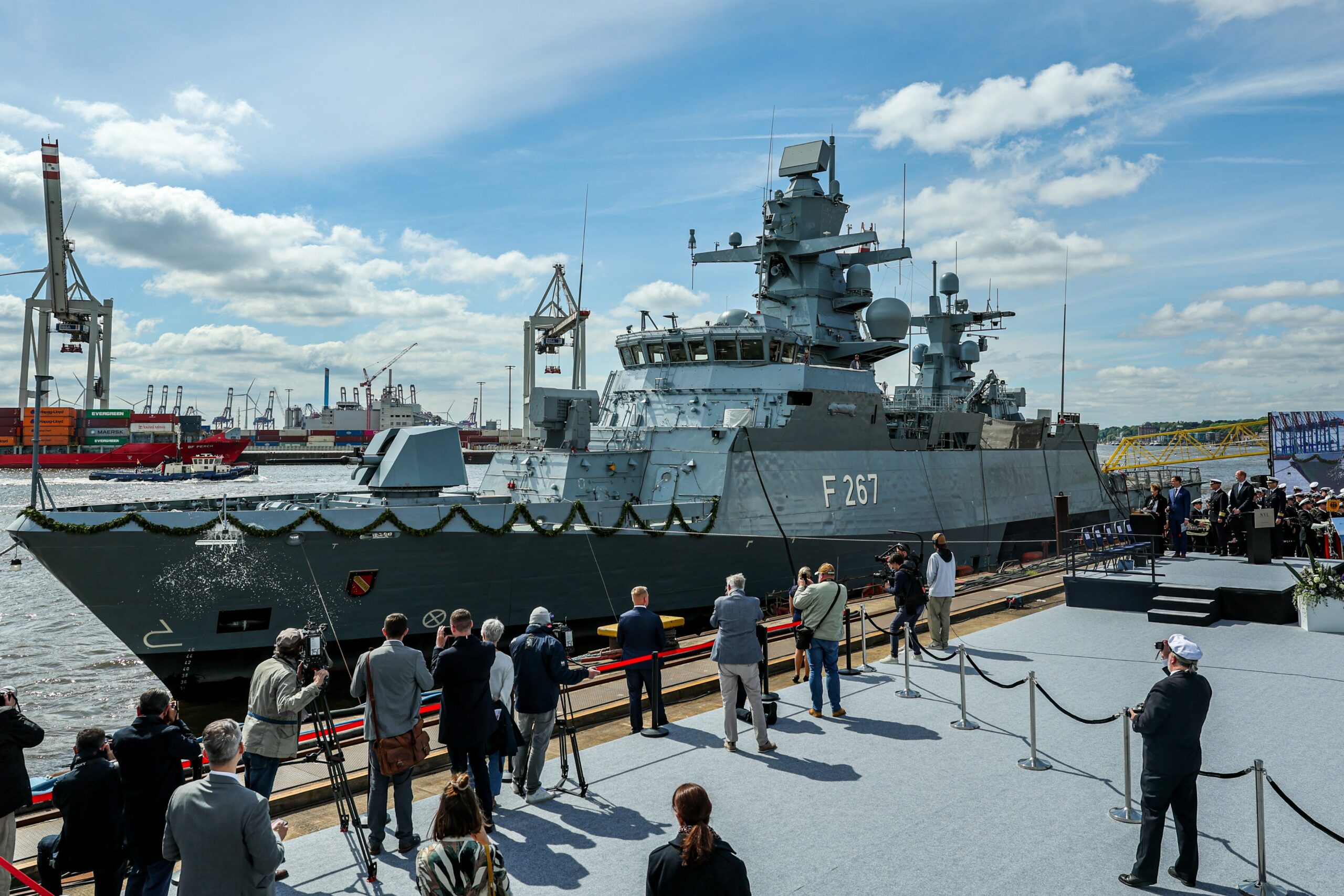 The Karlsruhe, a German Navy corvette, is seen docked in a pier during its christening ceremony. The gray ship's deck railings are decorated with a ribbon made of leaves. The middle of its hull has 