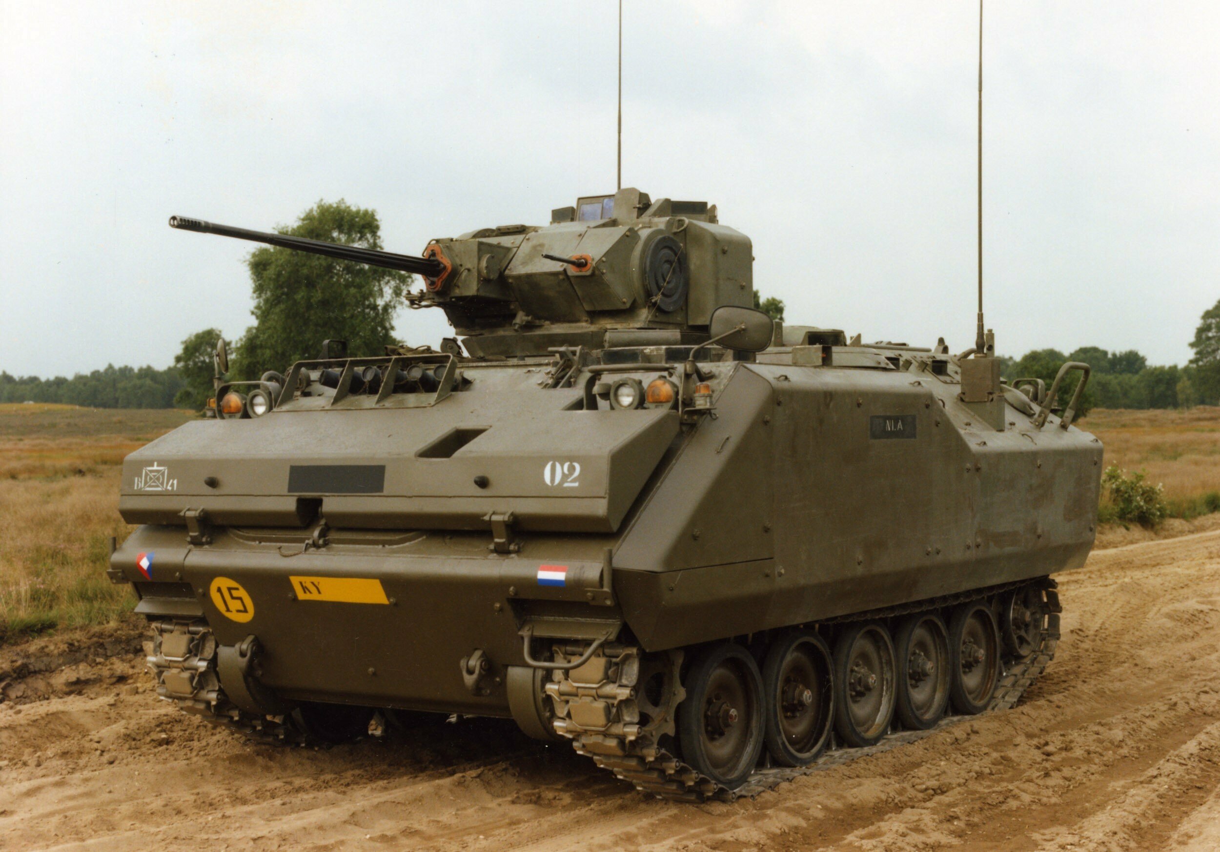 A YPR-765 armored infantry fighting vehicle is seen stationed on a dirt road. The vehicle has the typical appearance of a military tank, with a boxy built, a turret, and tracked wheels. The vehicle is painted grayish green. The background is a horizon of green trees and a clear white sky.