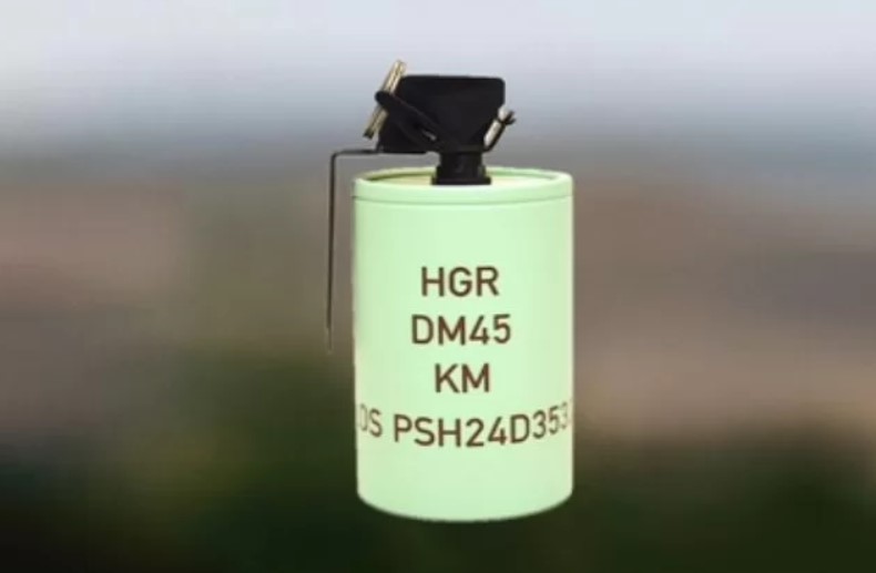 A Rheinmetall DM 45 hand grenade is displayed. The grenade has a cylindrical shape and is painted green, with 