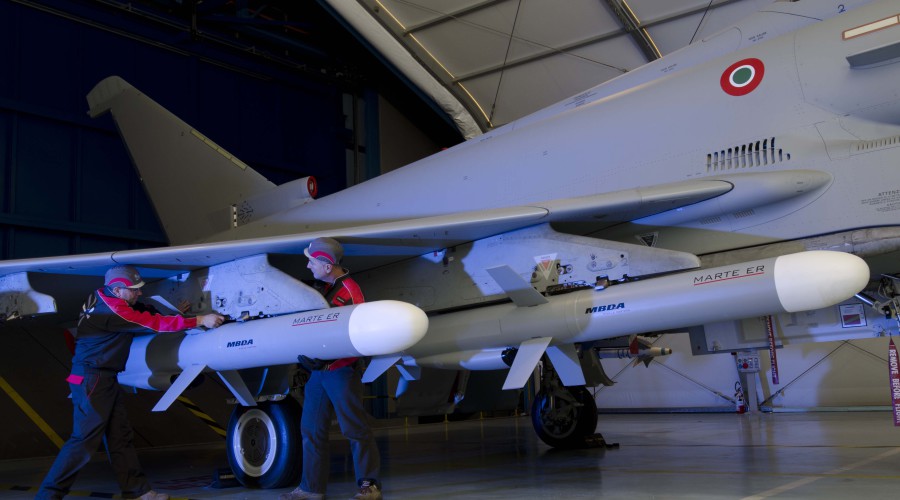 Two gray MBDA Marte ER missiles are seen attached to a similarly-painted jet plane parked inside what appears to be a hangar. Two workers inspect one of the missiles set underneath the plane's right wing.