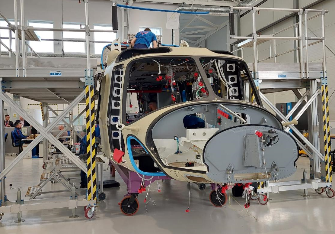 An AW149 Helicopter is seen in the middle of production. It has incomplete parts, but the shape of the front of the aircraft already resembles that of a usual helicopter. Wires and exposed framework are seen on the vehicle. A worker wearing a blue shirt is seen coming up out of its roof. Other personnel are seen in the far back in what appears to be an office space with desks and cubicles.