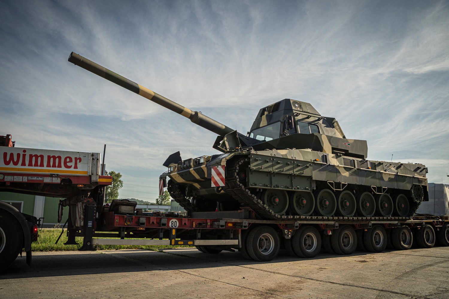 A Leopard 2A7HU battle tank is seen loaded on the back of a truck. The tank is painted in camouflage.