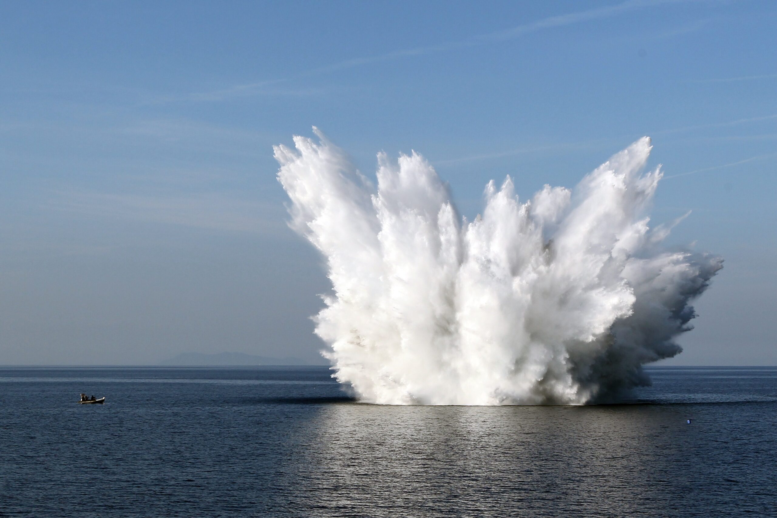 A naval mine sets off in the middle of an ocean. The water's white mist is seen blasting off on all directions. On the far left side of the image, a small boat is seen.