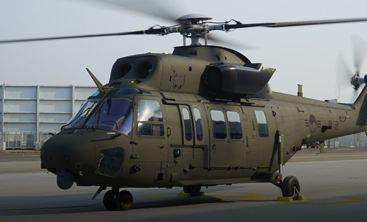 A KUH-1 Surion Helicopter is seen grounded in what appears to be a large open area, most likely a testing facility or a military headquarter. The aircraft is painted dark green, and its propellers and rotor blades are blurry as they spin.