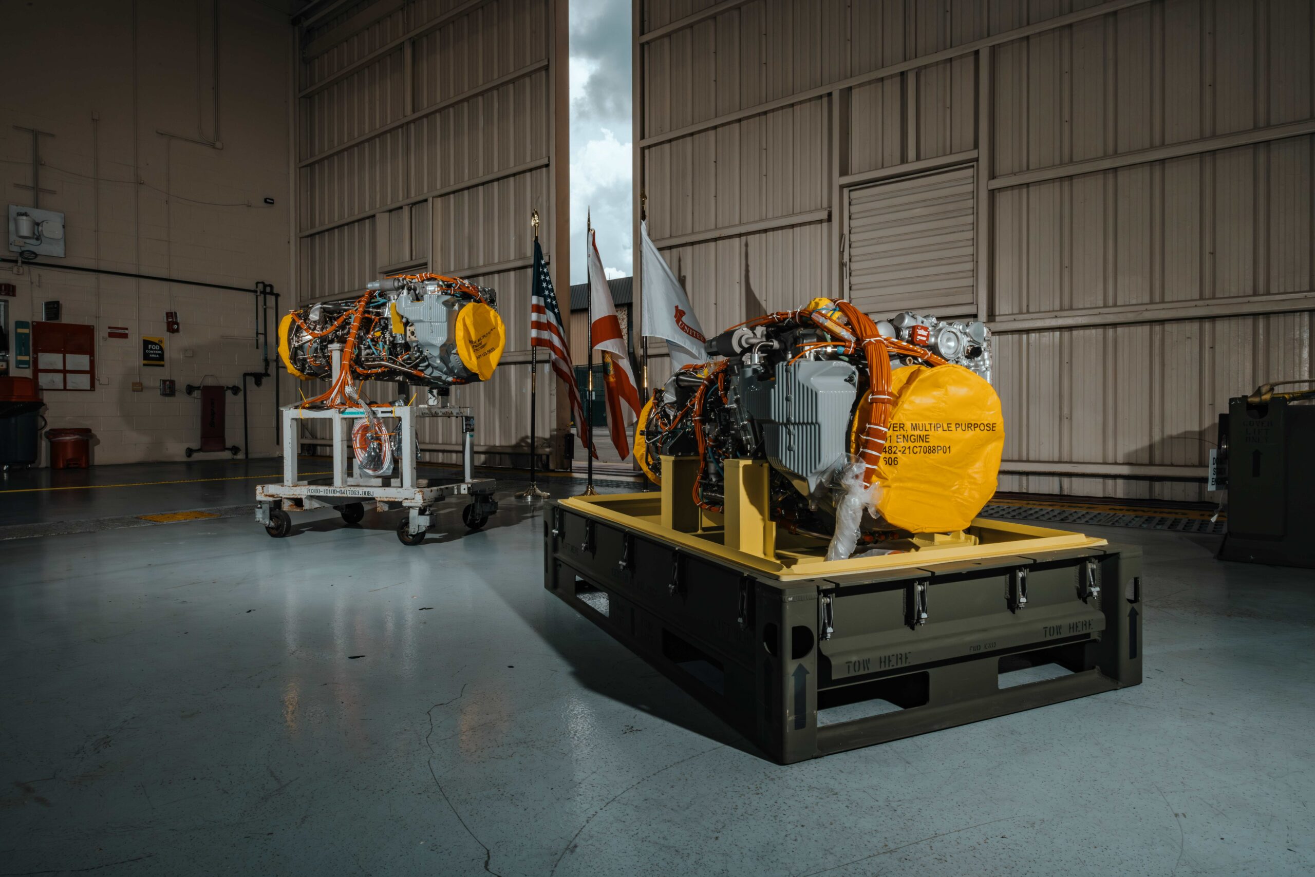Two General Electric (GE) Aerospace T901 engines are displayed inside what appears to be a big warehouse or aircraft hangar. Wires, exposed metal, and other parts of machinery make up the engines.