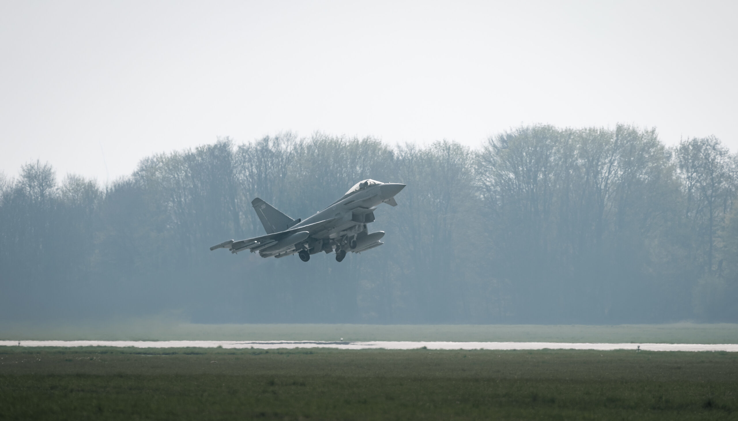 A UK Royal Air Force Typhoon jet takes off. The aircraft is colored gray, with 350 painted on its rudder. The background, partially covered in fog, appears to be a forested area.
