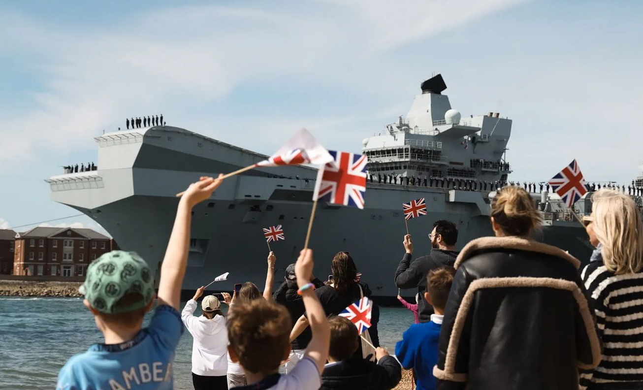 UK Royal Navy's HMS Prince of Wales is seen leaving Portsmouth Naval Base. 11 people bid farewell in the foreground, waving UK flags.