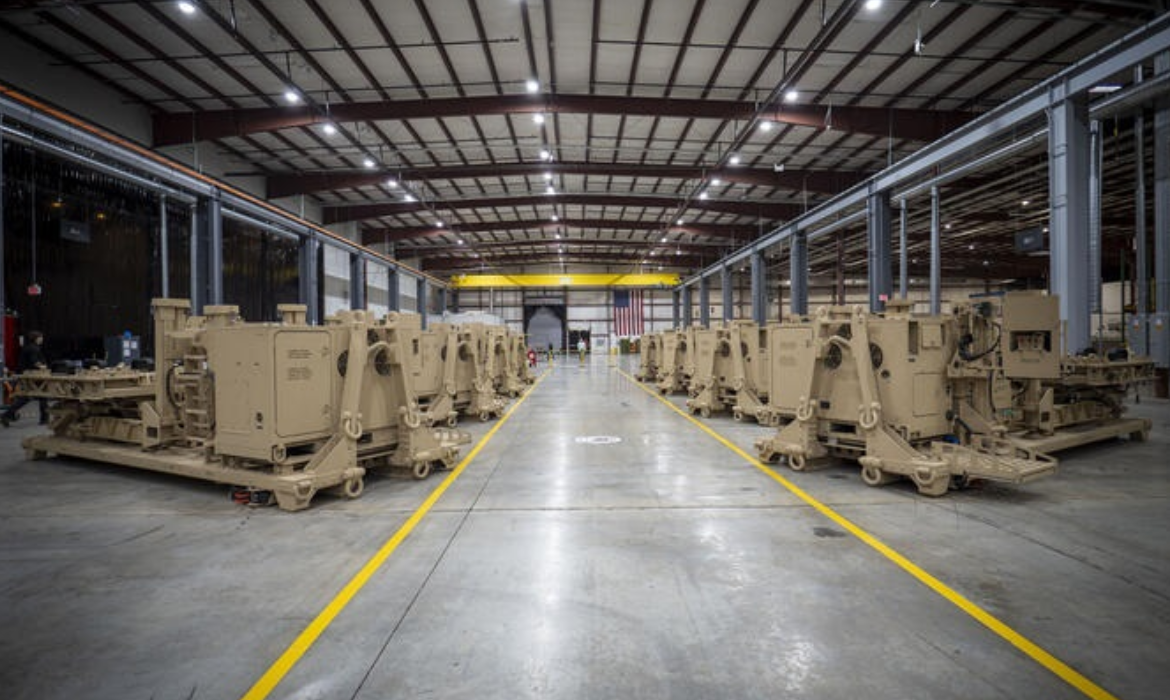 About eight units of Indirect Fires Protection Capability Increment 2 (IFPC Inc 2) ground-based systems are seen lined up in two rows in a large facility that appears to be a warehouse or a factory. The large units are colored light brown.