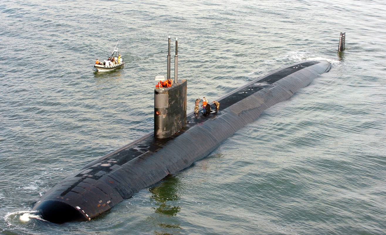 USS Toledo (SSN 769) is seen partially submerged in deep, blue-green waters. About six crewmen are seen atop the submarine. A small patrol boat is seen beside the submarine, on the upper left side of the image, with four people on board.