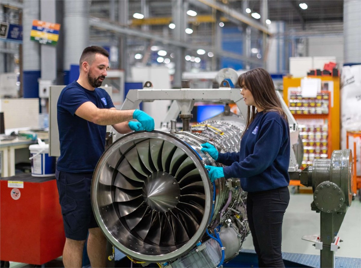 Two individuals wearing navy blue shirts with the Rolls-Royce logo on the left side of the chest are see inspecting a Typhoon aircraft engine. They appear to be inside an industrial building, with steel equipment and finishing in the blurred background.