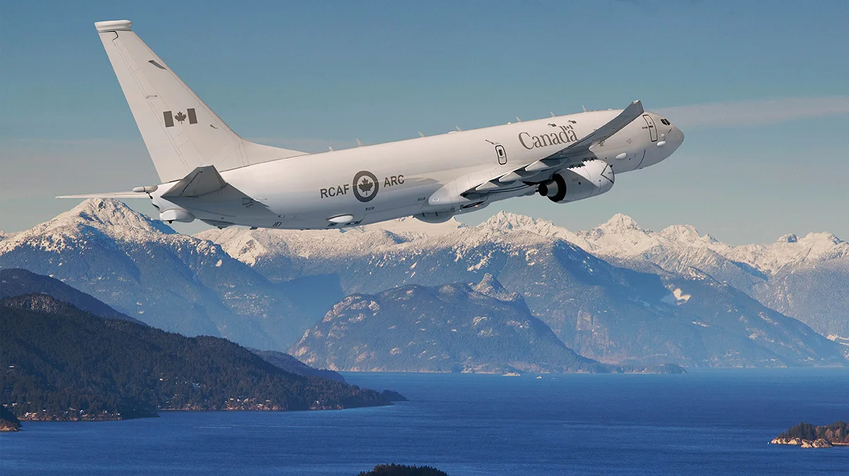 A Royal Canadian Air Force P-8A Poseidon aircraft is seen flynig over a large body of deep-blue water. In the far background, a snow-capped mountain range can be seen.