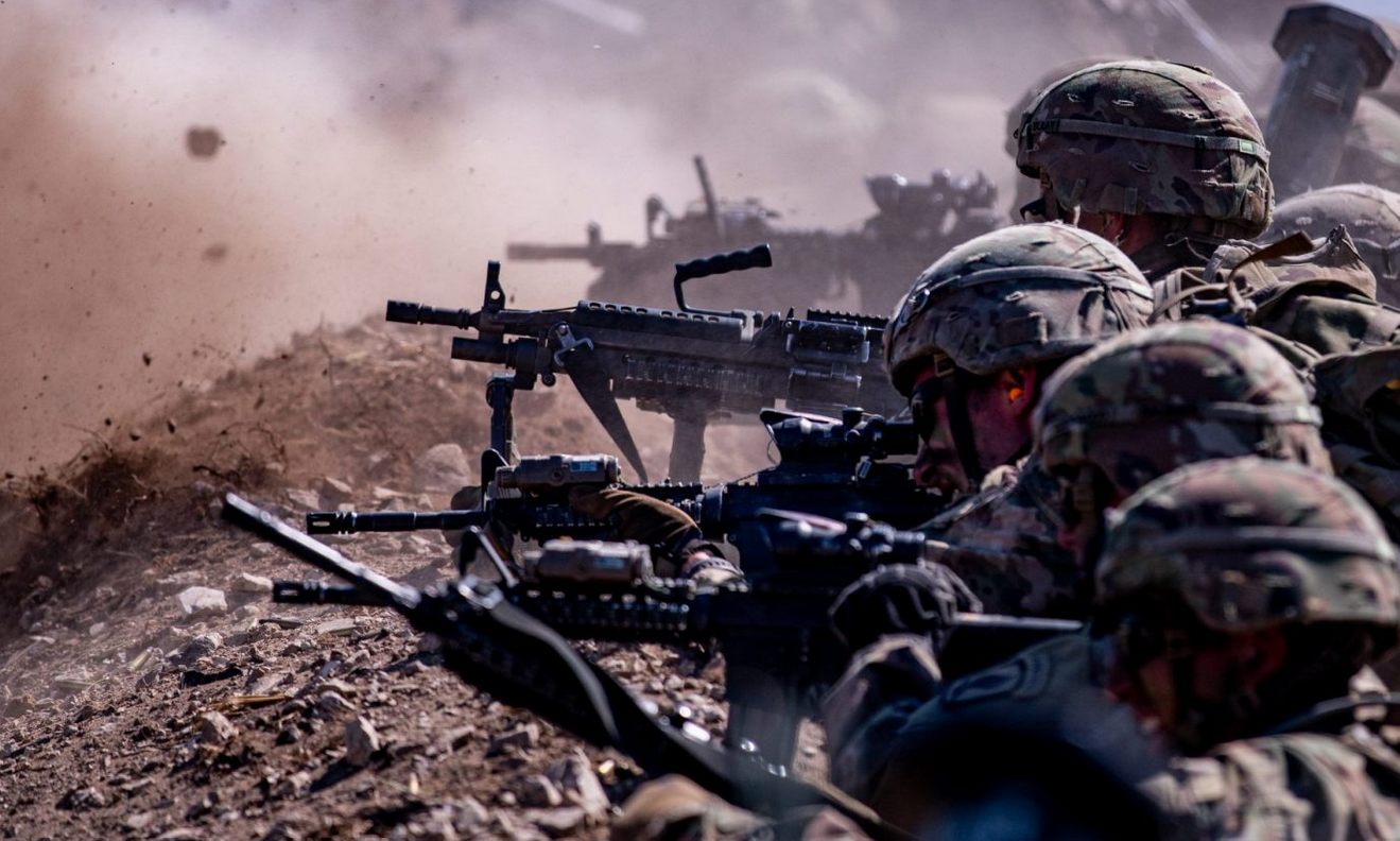 Four camo-wearing soldiers are seen positioned in a trench, pointing guns toward a target that is out of frame.