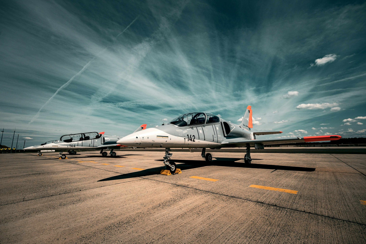 The Hungarian Air Force L-39 Skyfox aircraft is seen stationed on the ground in what appears to be a runway of an air base. The aircraft is painted gray on the upper half of its body, and white on the lower half and on its front. The body number 142 is painted black on its body's left side. The wings and tail's tips are painted a bright red-orange. On its far right, another Skyfox is seen parked. It has the number 144 painted on it.