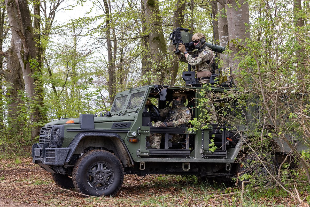 An ENOK AB light armored patrol vehicle is seen stationed in what appears to be a forest. The car is painted forest green, and two soldiers wearing green camouflage are seen crewing it. One is sitting in the driver's seat, while the other is on the back of the truck, aiming a high-millimeter rifle resting on his shoulder at something out of frame.