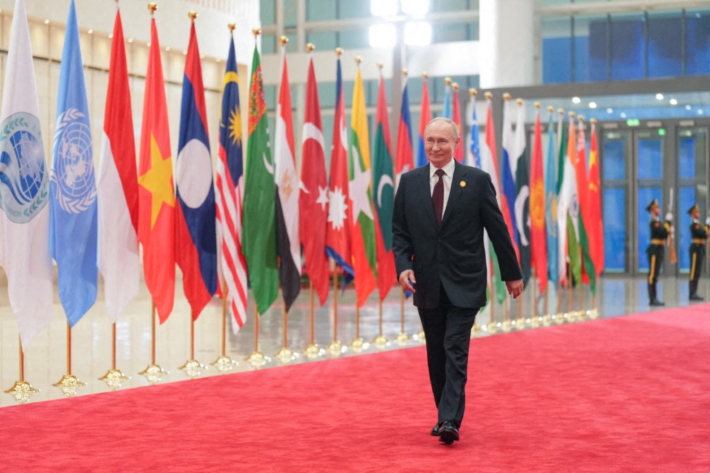 Vladimir Putin walking over a red carpet with country flags in the background
