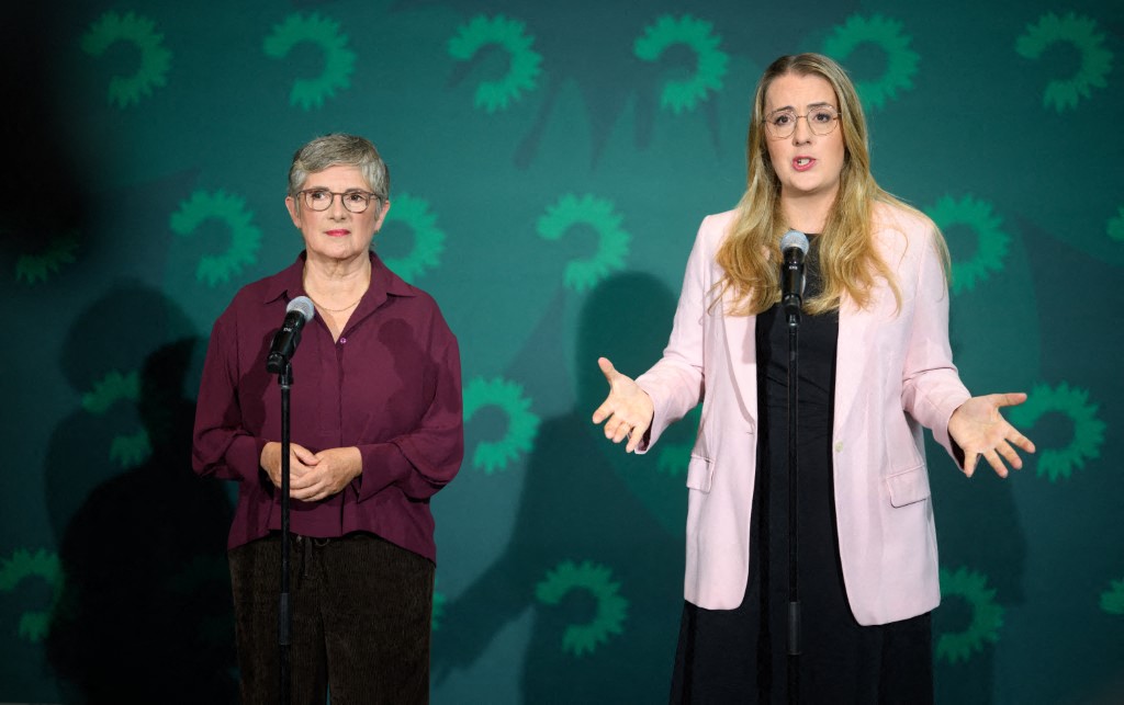 Britta Haßelmann wearing a maroon blouse, and Katharina Dröge wearing a light pink blazer over a black dress while speaking at a press conference with a green background