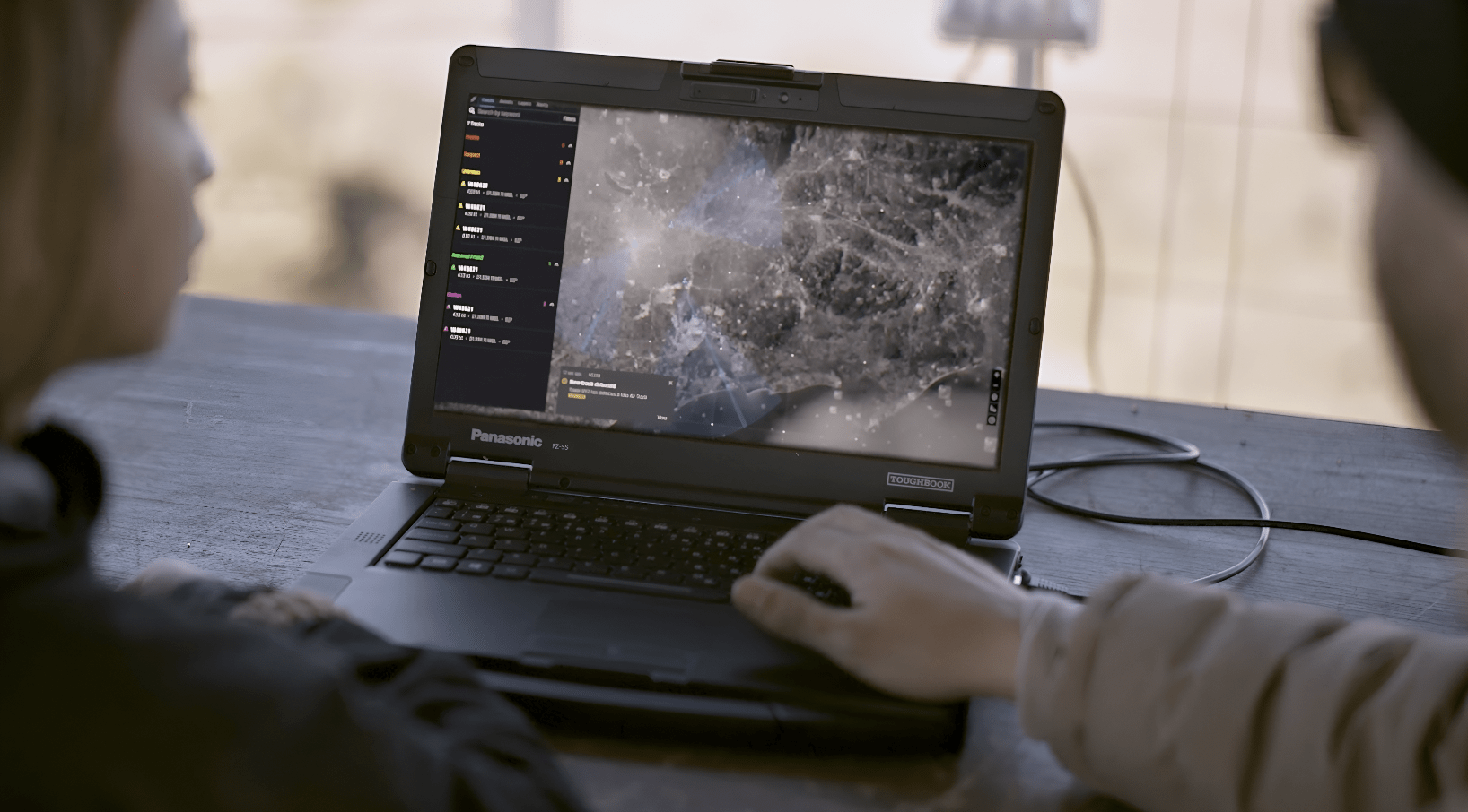 Defense operators using real-time drone surveillance software on a Panasonic Toughbook for mission control and target tracking.