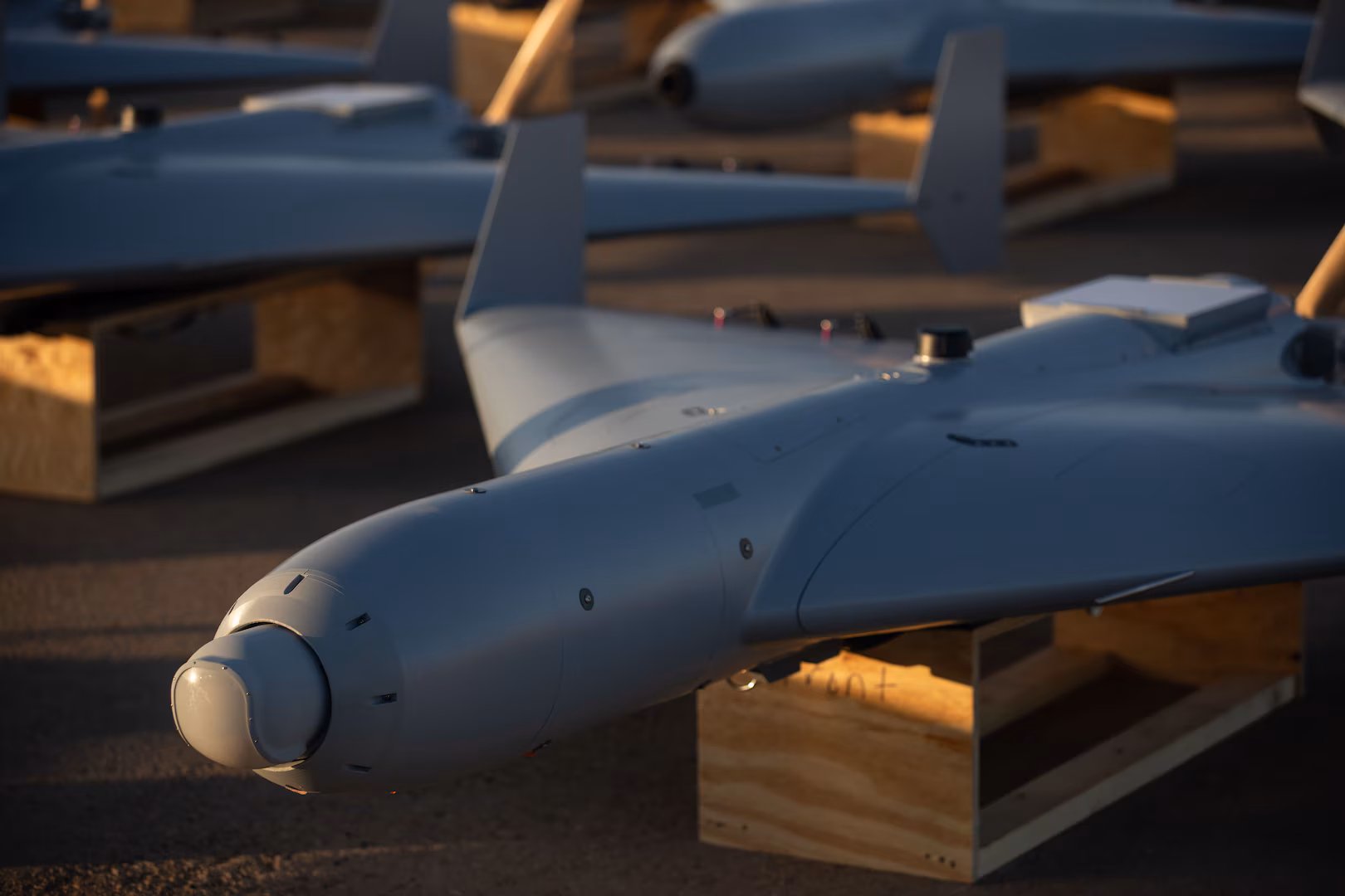 Close-up view of a LUCAS drone resting on a wooden stand, showing its sleek fixed-wing design and nose-mounted sensor under warm evening light.