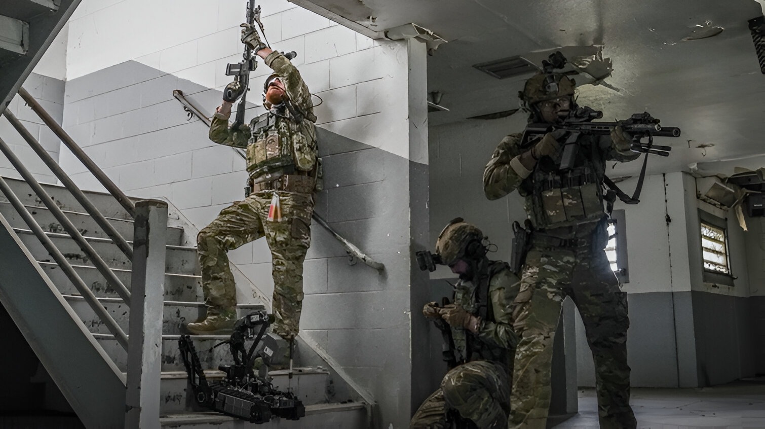Soldiers conduct training inside a damaged building, moving up a stairwell with rifles raised while an AeroVironment ground robot supports their indoor clearing exercise.
