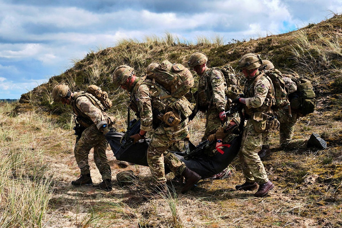 UK soldiers in full combat gear carry a casualty on a stretcher across grassy terrain during a blast-response training scenario