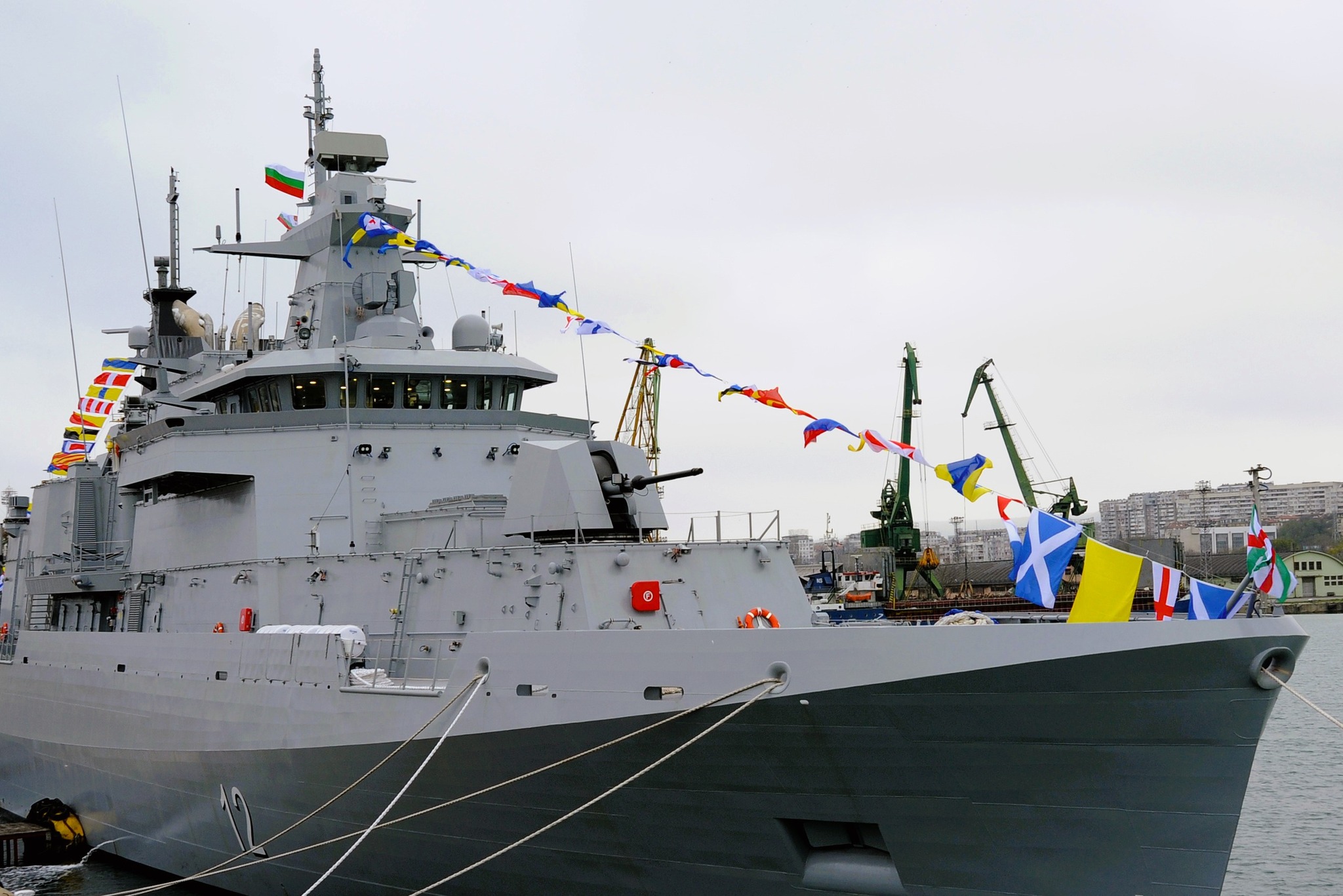 A modern grey Bulgaria warship docked at a harbor, decorated with colorful ceremonial naval flags and featuring a visible gun turret and radar mast against a cloudy sky