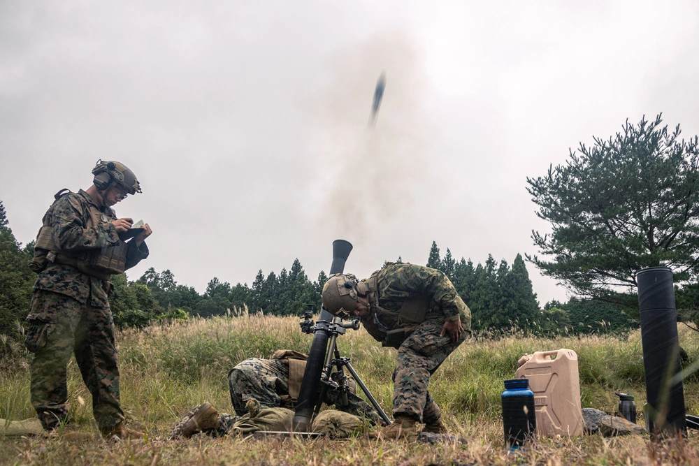 Soldiers conduct a live mortars firing during a field exercise, with one round captured mid-launch from the tube as the crew coordinates targeting and safety procedures in a grassy training area
