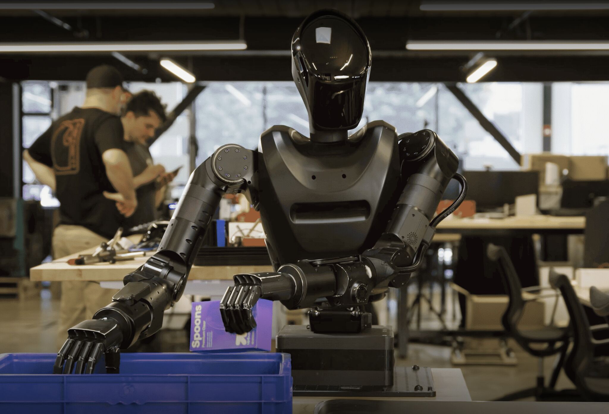 Humanoid robot operating in a robotics lab, using articulated arms to handle objects while engineers work in the background
