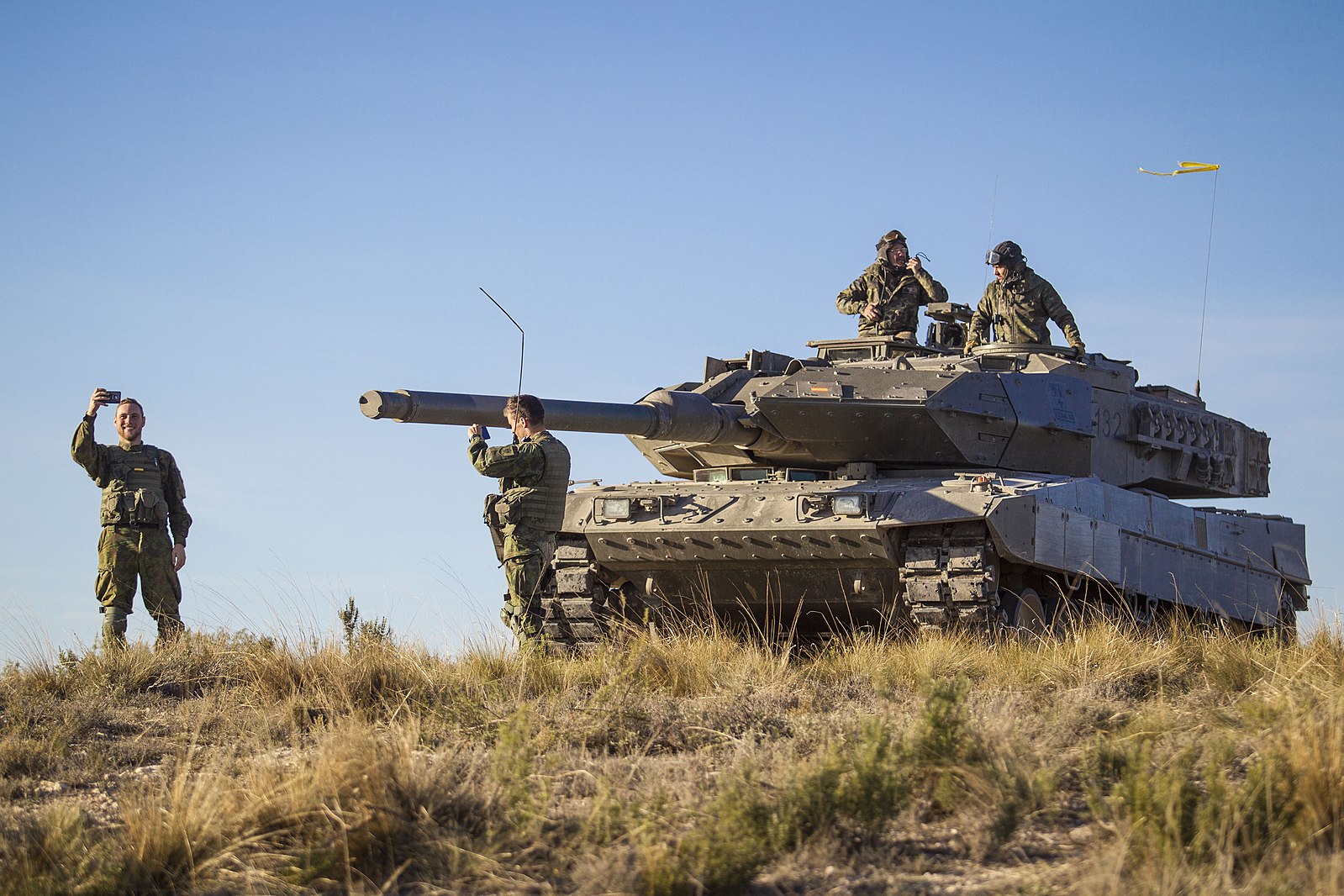 Soldiers stand and communicate beside a Leopard battle tank positioned on open terrain during daylight, with crew members visible on the turret and the main gun facing forward under a clear sky