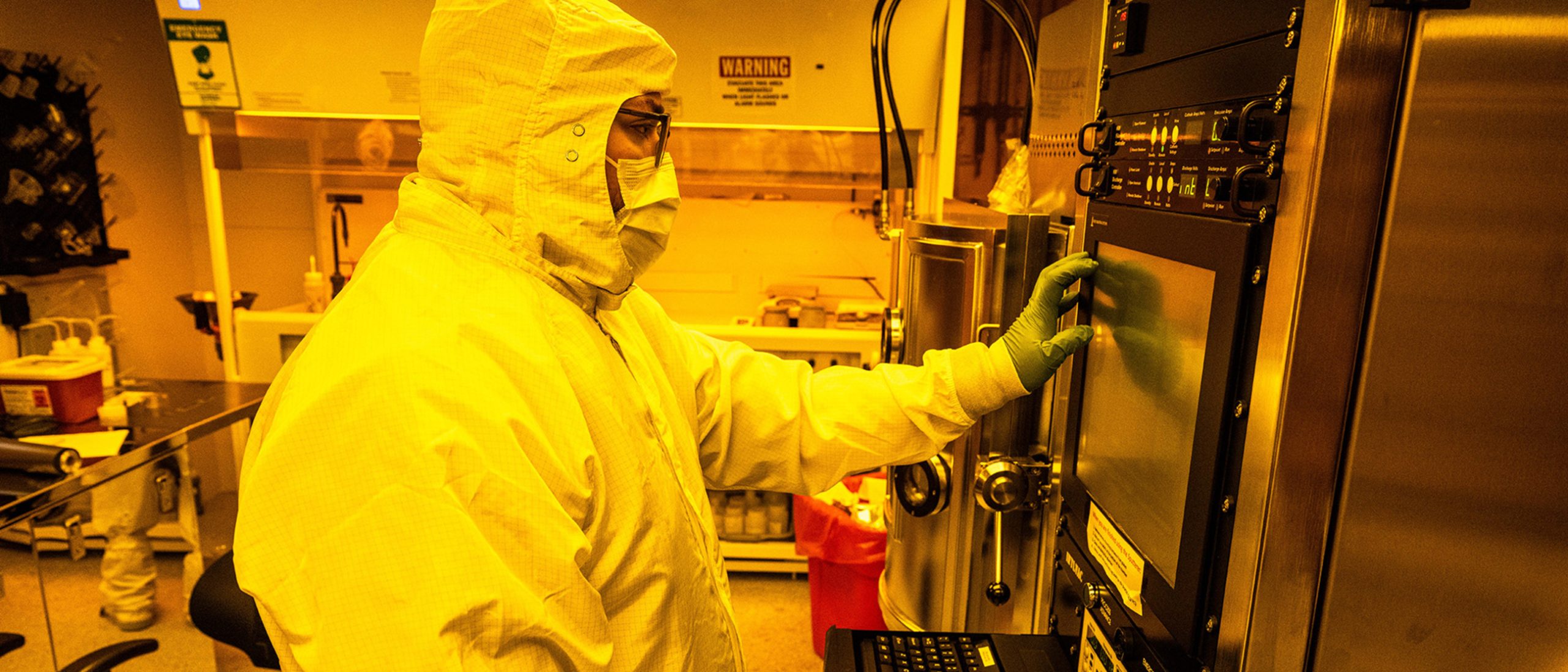 Technician wearing full cleanroom protective gear operates advanced quantum fabrication equipment inside a yellow-lit semiconductor cleanroom, interacting with a control panel on a precision processing system
