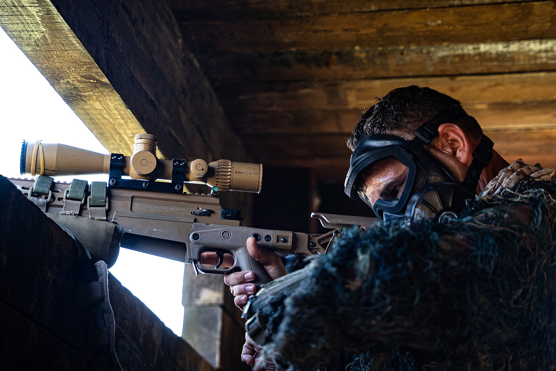 Soldier wearing protective goggles aims a sniper from inside a concrete fighting position during a military training exercise.