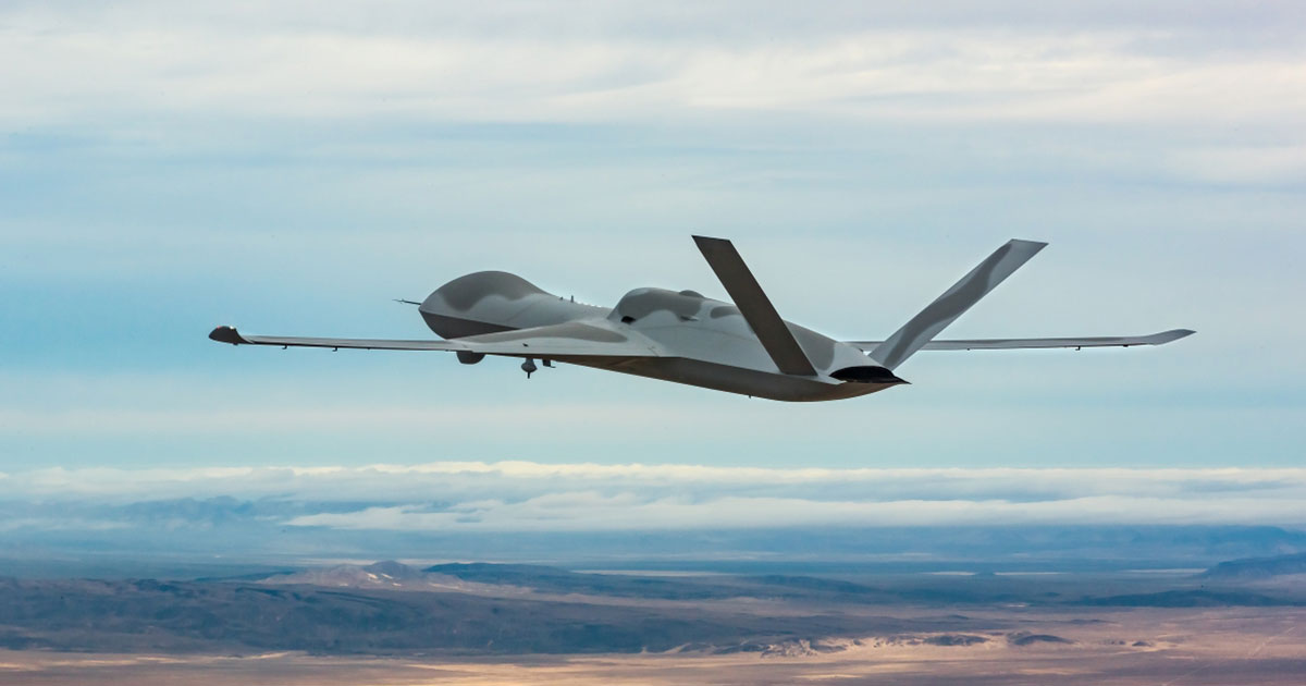 MQ-20 Avenger drone flying at altitude over a desert landscape, showing its stealthy blended-wing design, rear-mounted engine, and long wingspan during an autonomous flight demonstration.