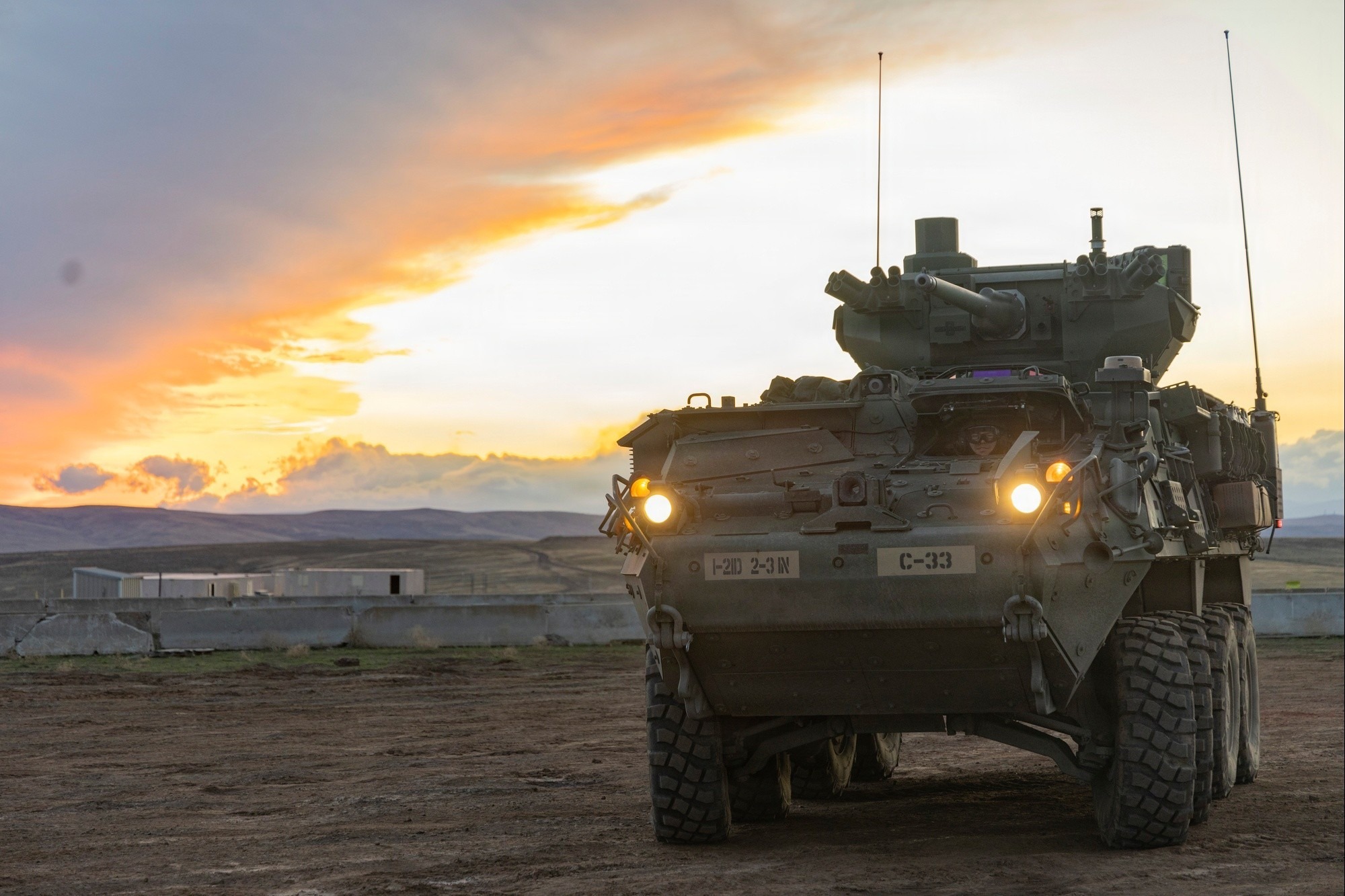 Stryker armored combat vehicle positioned on a dirt training range at sunset, headlights illuminated, with its remote weapon station visible against an open landscape.