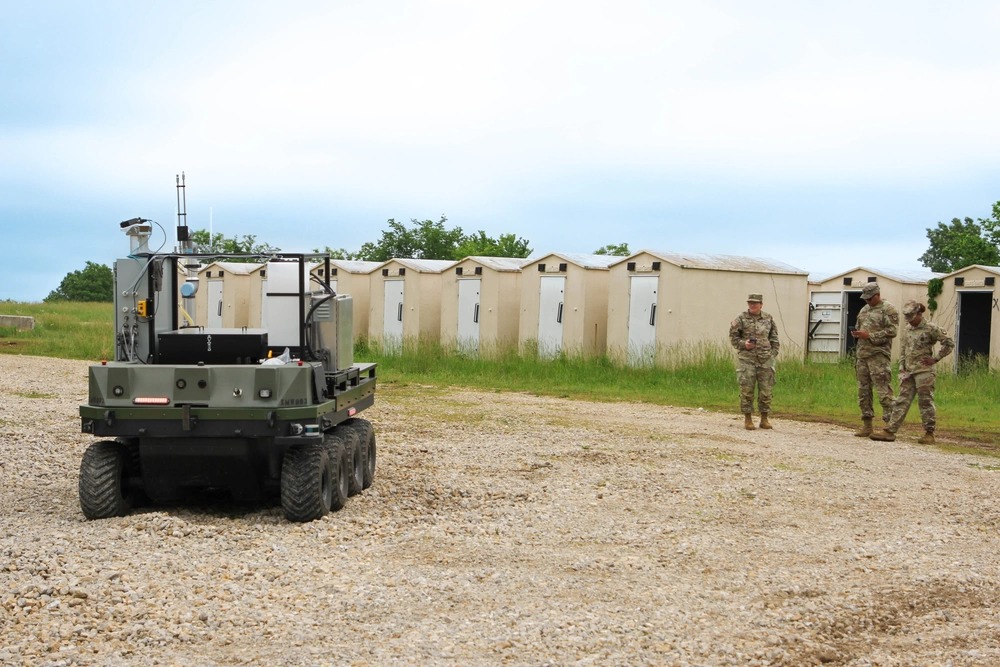 Military personnel observe autonomous ground robots operating in a field near temporary structures during a training or testing exercise.
