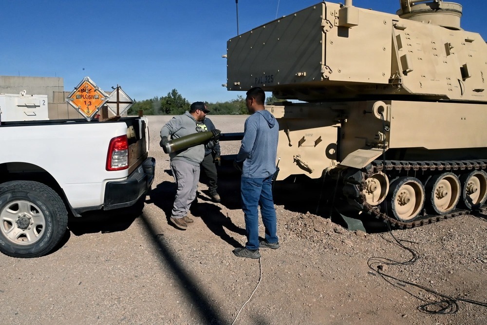 Personnel load an artillery round beside a tracked armored vehicle at a training site, with equipment and support vehicles nearby.