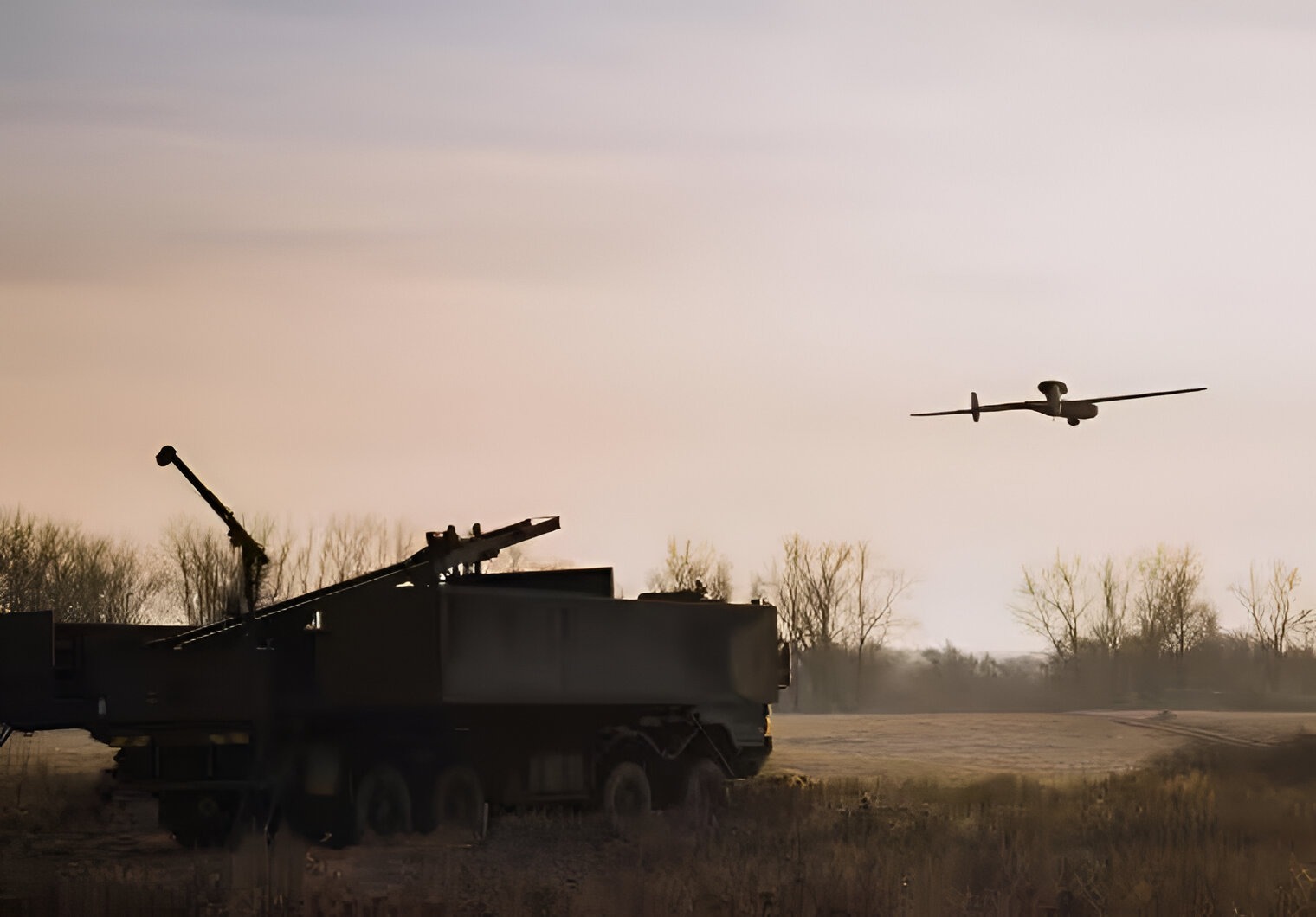 Rheinmetall’s LUNA NG unmanned aerial vehicle flying during a Bundeswehr field trial, operating alongside a ground-based air defense system in a rural training area at dusk