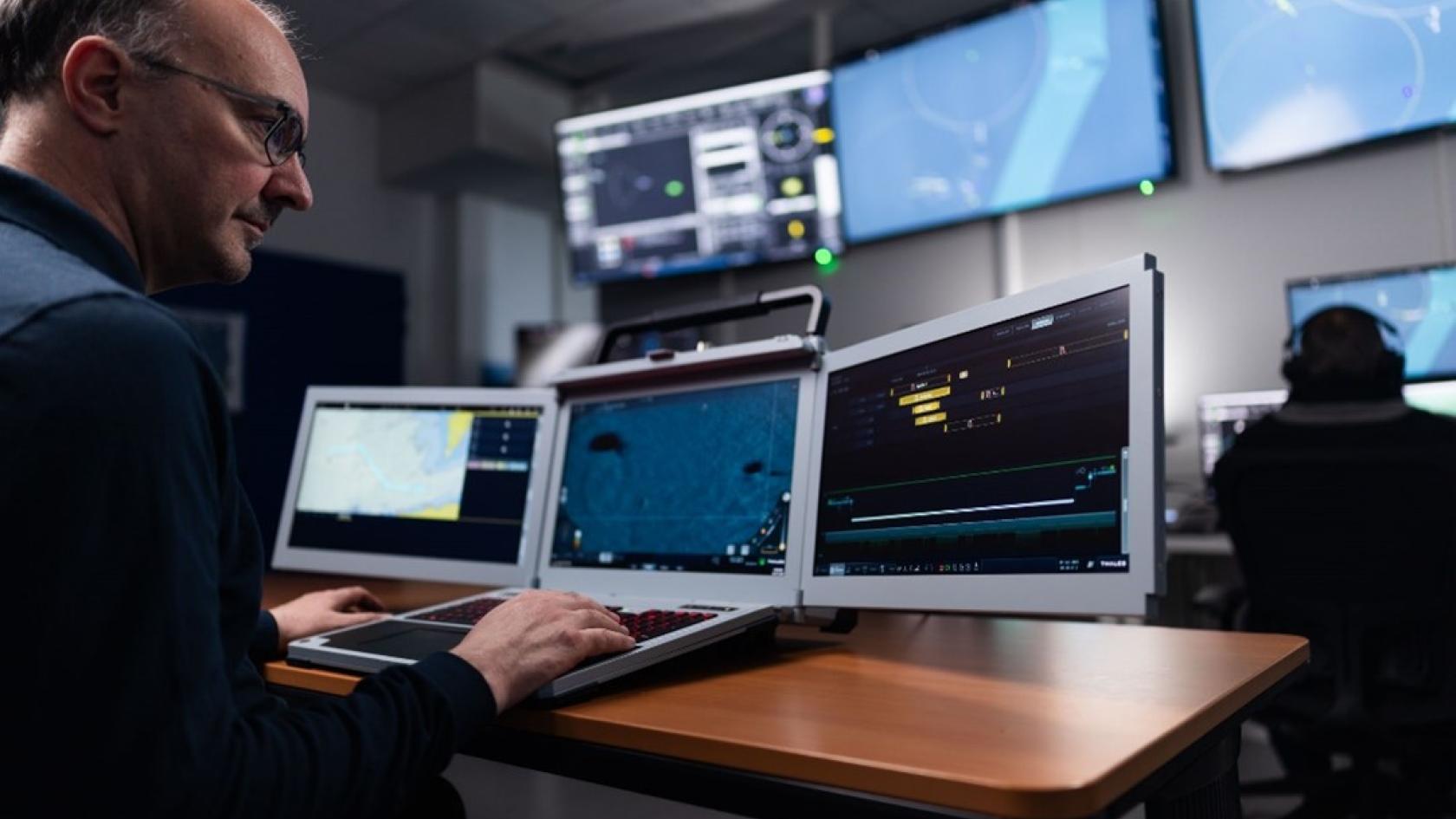 Thales technician operating a tri-screen laptop workstation displaying maritime surveillance, tracking, and tactical data feeds in a dimly lit defense operations center