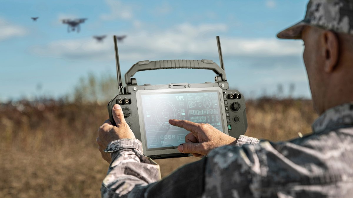 Soldier operating a Getac UX10 rugged ground control station touchscreen in an open field with multiple military drones visible in the background, illustrating a modular unmanned systems command center