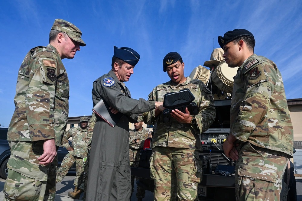 A US Air Force pilot officer and several airmen in OCP uniforms huddle around a handheld tactical device near field equipment, illustrating collaborative efforts to solve emerging drone warfare challenges