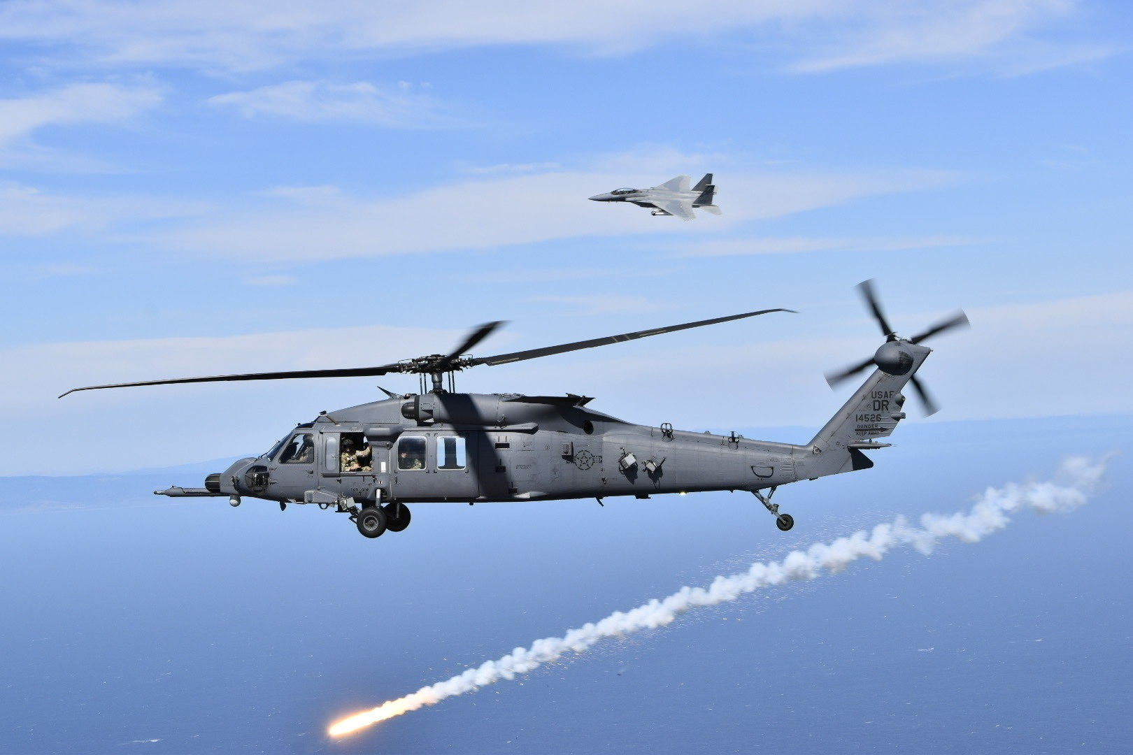 U.S. Air Force HH-60W Jolly Green II assigned to the 305th Rescue Squadron, Davis Monthan Air Force Base, Ariz., deploys countermeasure flares as an U.S. Air Force F-15D Eagle from the 144th Fighter Wing, California Air National Guard, maneuvers overhead during a joint over-water test flight, Aug. 20, 2025. (U.S. Air Force courtesy photo)