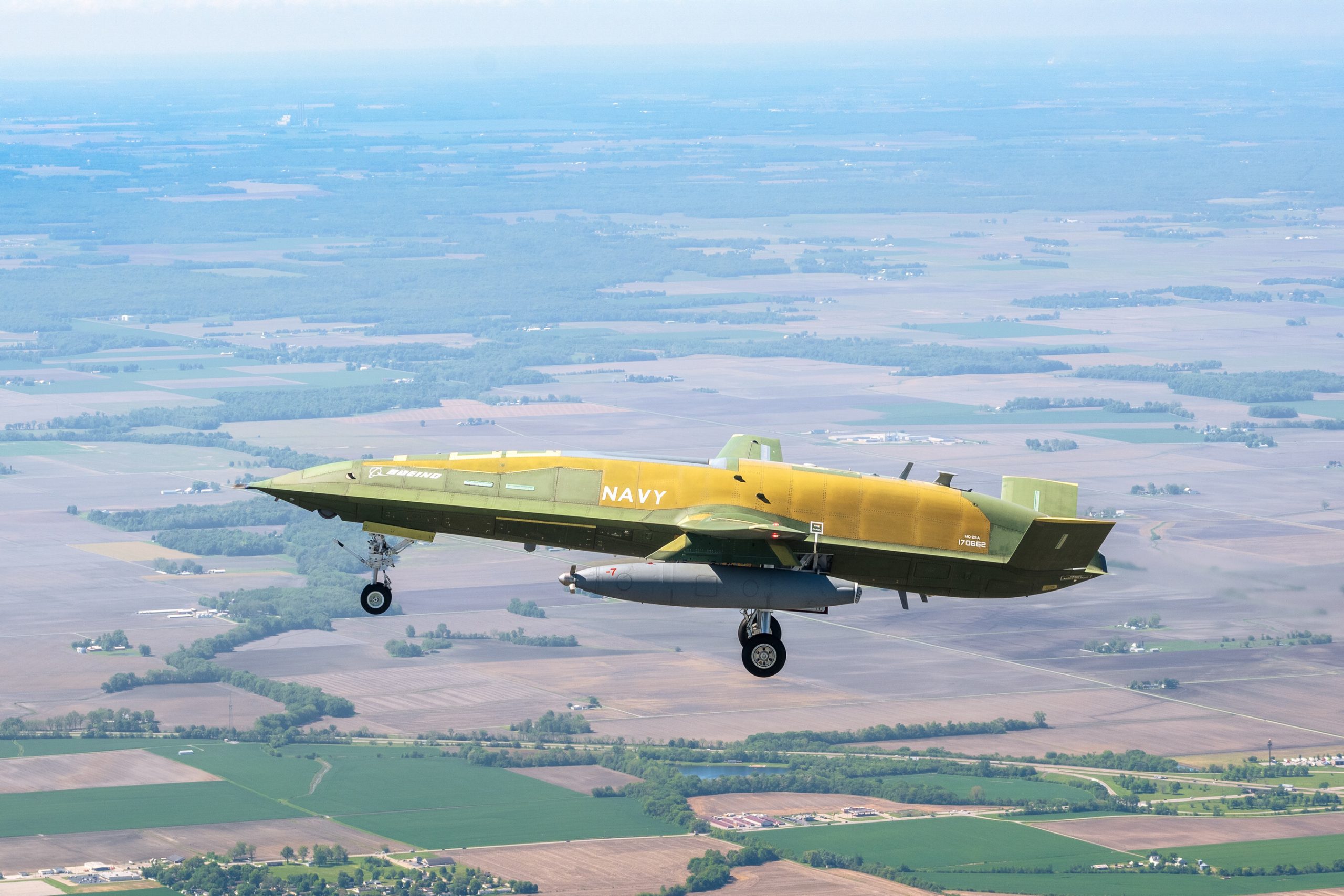 Boeing MQ-25A Stingray unmanned tanker aircraft in green primer and yellow livery during its historic first flight over a rural landscape for the U.S. Navy