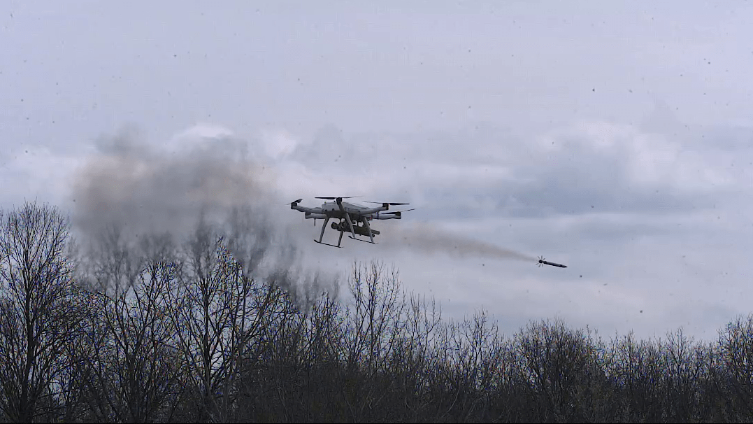 Serbian military autonomous quadcopter drone firing an aircraft rocket with a smoke plume trailing behind it over a bare winter treeline, showcasing the platform's multi-rocket air-to-ground strike capability